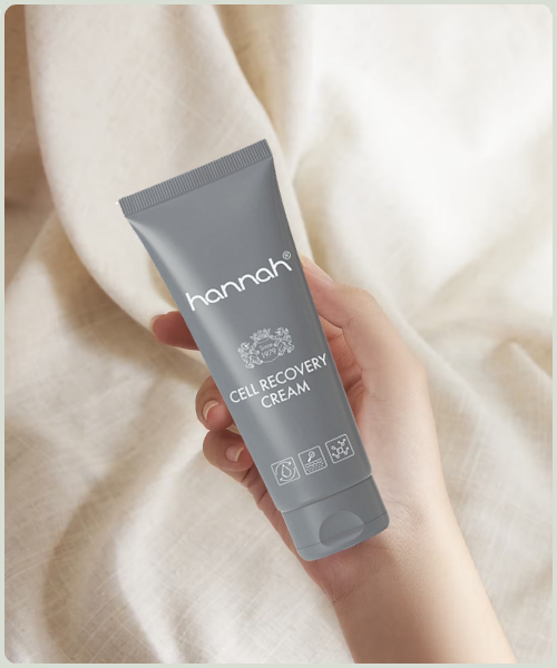 Een hand die een tube hannah Cell Recovery Cream vasthoudt tegen een achtergrond van zachte, lichte stof.