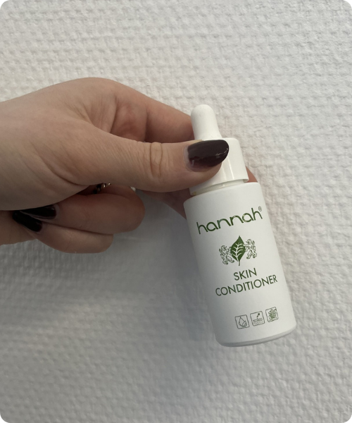 Een hand met donker gelakte nagels houdt een wit flesje hannah Skin Conditioner vast tegen een witte, gestructureerde achtergrond.