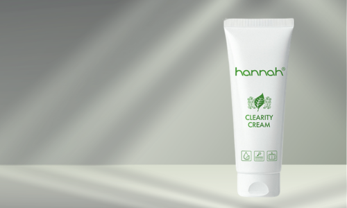 Een witte tube van hannah Clearity Cream met groene letters en logo, staand op een lichte ondergrond tegen een achtergrond met zachte lichtinval.