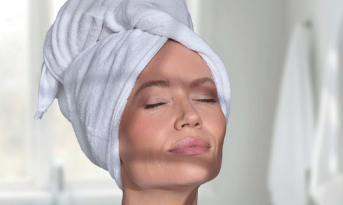 Een vrouw met een witte handdoek om haar hoofd en schouders, met gesloten ogen genietend van een moment van rust.
