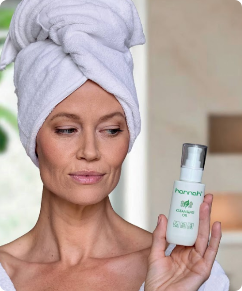 Vrouw met een handdoek om haar hoofd die een flesje hannah cleansing oil vasthoudt.