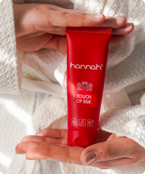 Een rode tube hannah Touch of Silk huidverzorgingsproduct geplaatst op een glanzende, champagnekleurige zijden achtergrond.