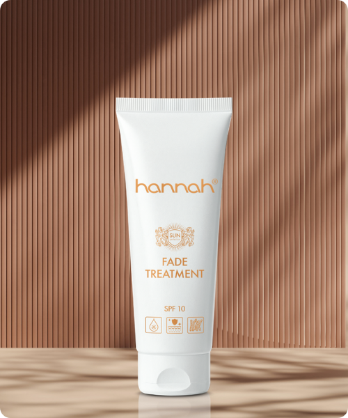 Een witte tube hannah Fade Treatment SPF10, geplaatst voor een achtergrond met een lijnenspel van licht en schaduw in warme bruintinten.