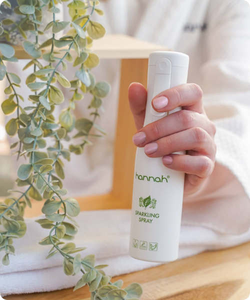 Een hand met roze nagellak die een witte flacon van hannah Sparkling Spray vasthoudt met op de achtergrond een witte badjas en hangende plant