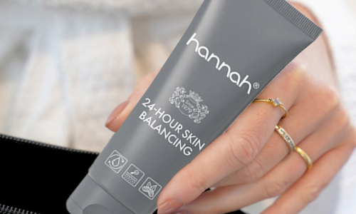 Een close-up van een hand met gouden ringen die een grijze tube hannah 24-hour Skin Balancing vasthoudt boven een zwarte tas.