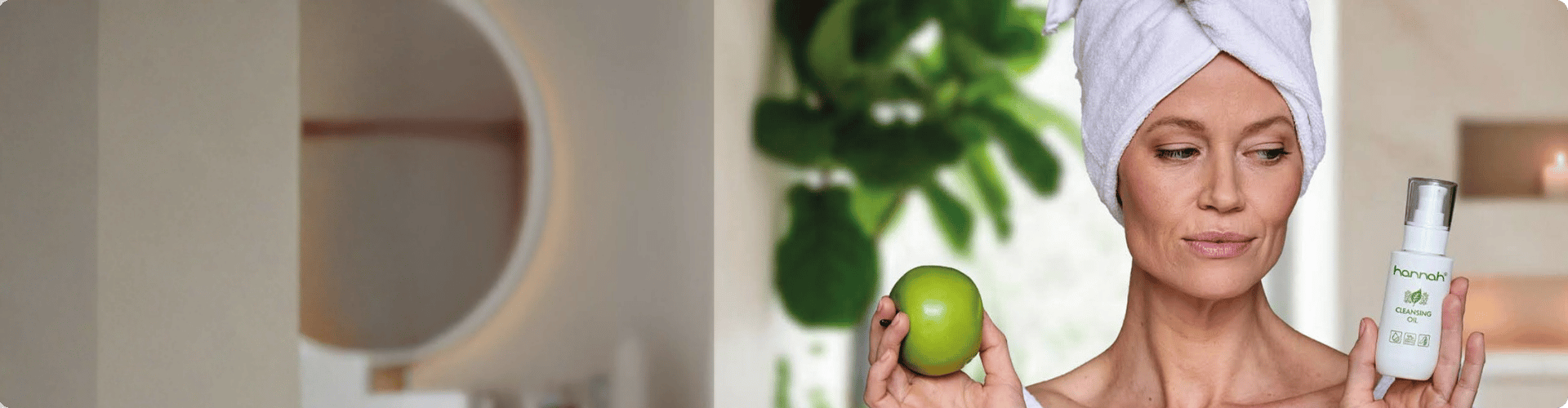 Vrouw met een handdoek op haar hoofd die een groene appel en een flesje hannah Cleansing Oil vasthoudt