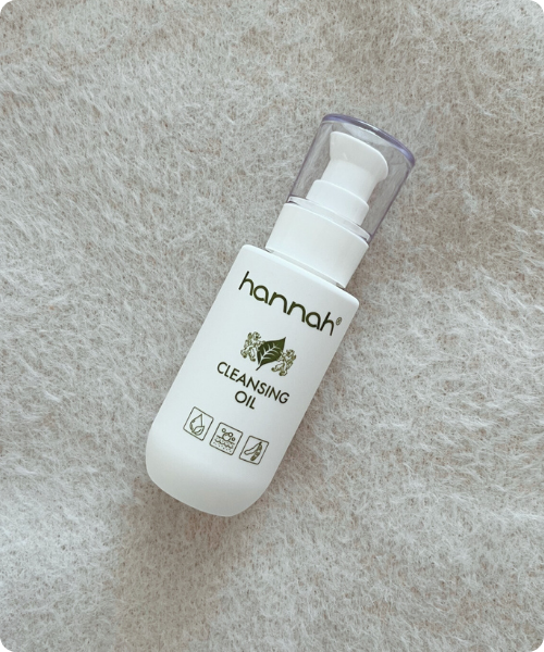 Een witte flacon van hannah Cleansing Oil met een handig pompje, gefotografeerd op een zachte, grijze ondergrond van imitatiebont.