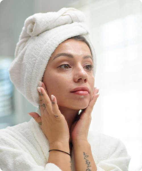 Een close-up van een vrouw in een witte badjas en met een handdoek om haar hoofd, die met beide handen de hannah Oxys Foros crème in haar gezicht inmasseert.