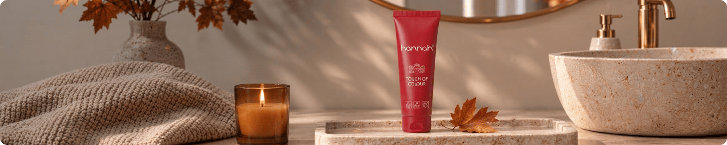 Een rode tube hannah Touch of Colour huidverzorging staat op een tray in een warme badkamer met een brandende kaars en herfstbladeren