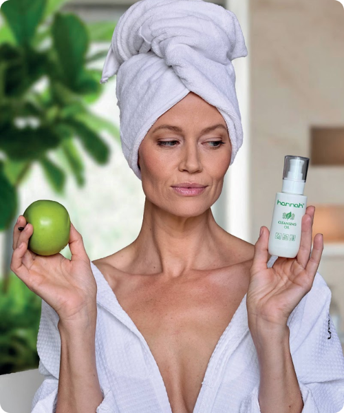 Een vrouw met een witte handdoek om haar hoofd houdt een groene appel en een flacon hannah Cleansing Oil vast.