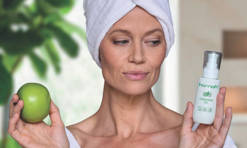 Een vrouw met een witte handdoek om haar hoofd houdt in haar ene hand een groene appel en in haar andere hand een flacon hannah Cleansing Oil vast.