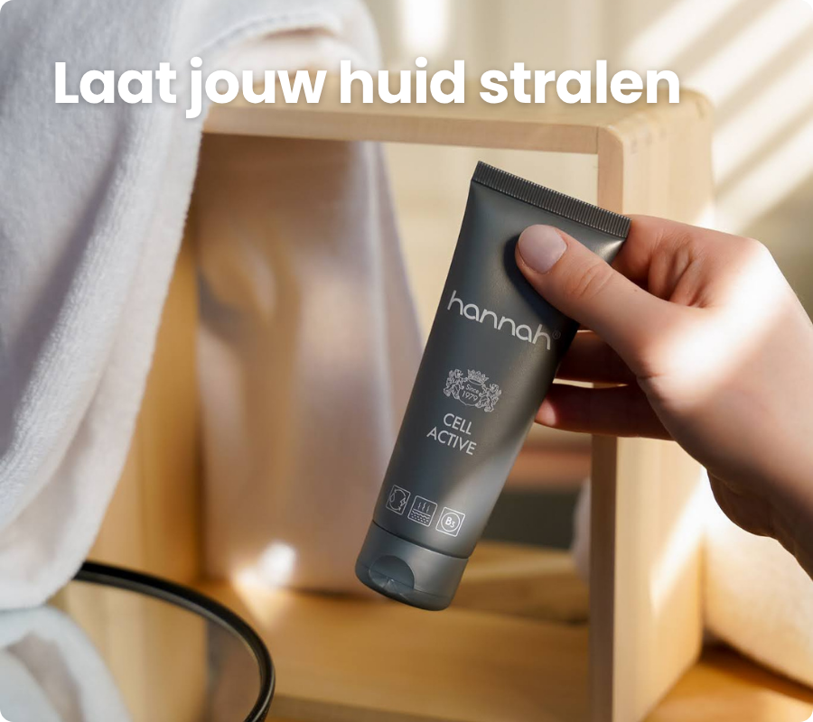 Een hand houdt een grijze tube hannah cell active vast voor een houten rek met een witte handdoek en een spiegel, met de nederlandse tekst Laat jouw huid stralen erboven.