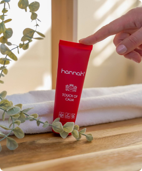 Een hand die met een vinger de bovenkant aanraakt van een rode tube hannah Touch of Calm naast een witte handdoek en groene plant