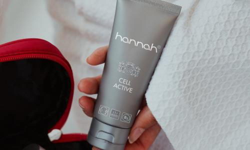 Een hand die een tube hannah Cell Active crème vasthoudt, met zichtbare iconen voor ingrediënten en huidvoordelen op de verpakking.
