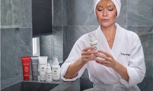 Een vrouw in een witte badjas en een haarband op haar hoofd bekijkt een flesje van het merk hannah in een badkamer, met een reeks andere hannah-producten naast haar op de wastafel.