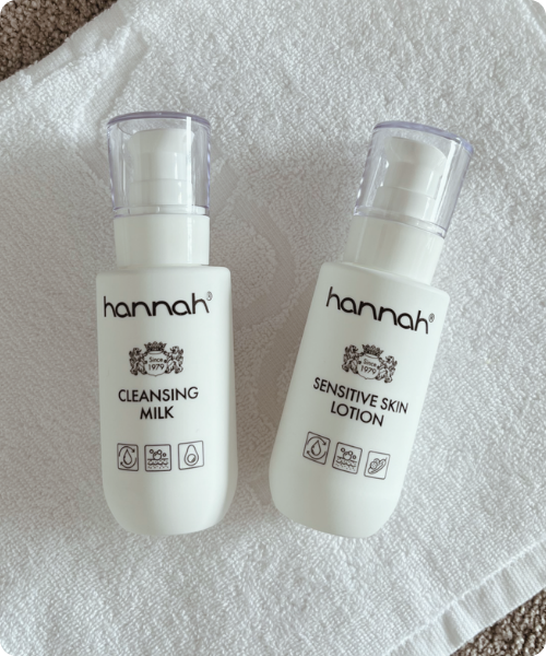 Twee witte flacons van het merk hannah, een Cleansing Milk en een Sensitive Skin Lotion, geplaatst op een witte kompresdoek.