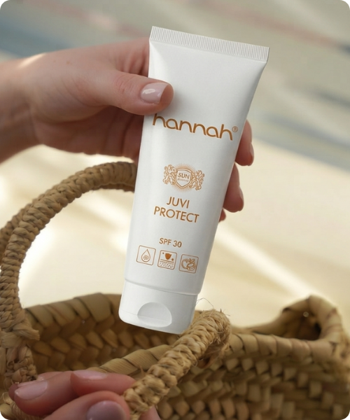 een hand die een tube hannah juvi protect spf 30 zonbescherming vasthoudt boven een gevlochten tas