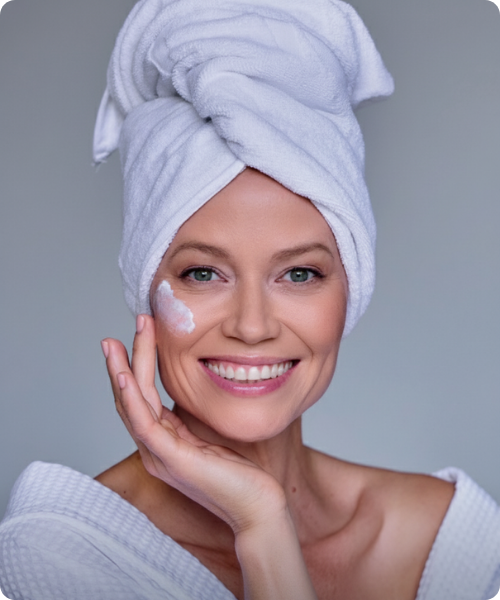 Vrouw met een handdoek om haar hoofd die een peeling product aanbrengt op haar gezicht.