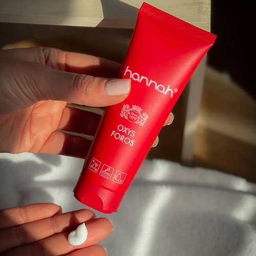 Een hand houdt een rode tube van het merk hannah vast met daarop de tekst Oxys Foros en in de andere hand ligt een beetje witte creme
