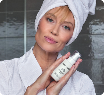 Een vrouw met een handdoek om haar hoofd houdt een flacon hannah cleansing milk vast voor een grijze tegelwand