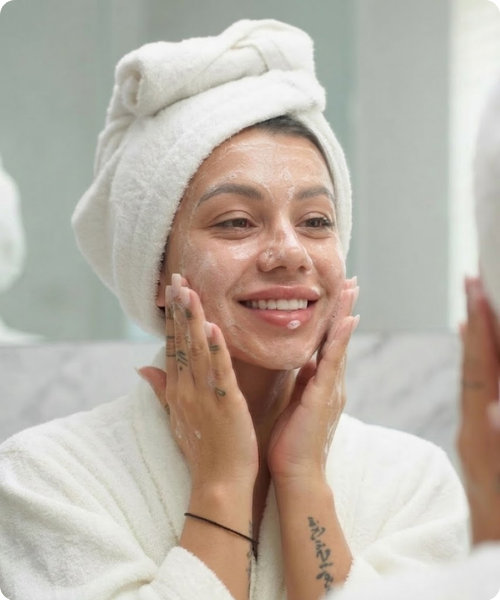 Een close-up van een lachende vrouw in een badjas met een handdoek om haar hoofd, die met schuim of crème op haar gezicht haar huid masseert voor de spiegel.