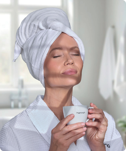 Vrouw met een handdoek op haar hoofd houdt ontspannen een wit hannah kopje vast in een badkamer.
