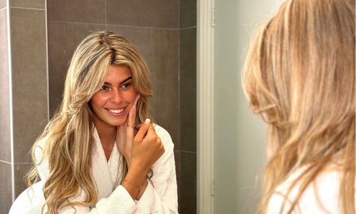 Een glimlachende vrouw met lang blond haar in een witte badjas, kijkend in de spiegel terwijl ze haar gezicht aanraakt na het gebruik van huidverzorgingsproducten.