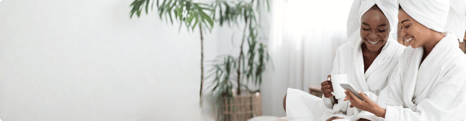 Twee lachende vrouwen in witte badjassen met handdoeken om hun haar kijken samen op een smartphone in een lichte kamer met een grote groene plant.