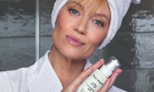 Een vrouw met een witte handdoek om haar hoofd houdt een flacon hannah Cleansing Milk vast tegen een grijze achtergrond.